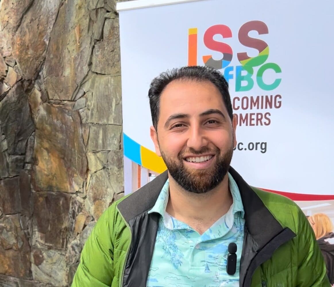 Former student of the LINC Squamish program, Joudi Saffo, smiling for the camera, wearing a jacket and t-shirt with a microphone attached to his clothes and a banner advertising ISSofBC's services behind him.