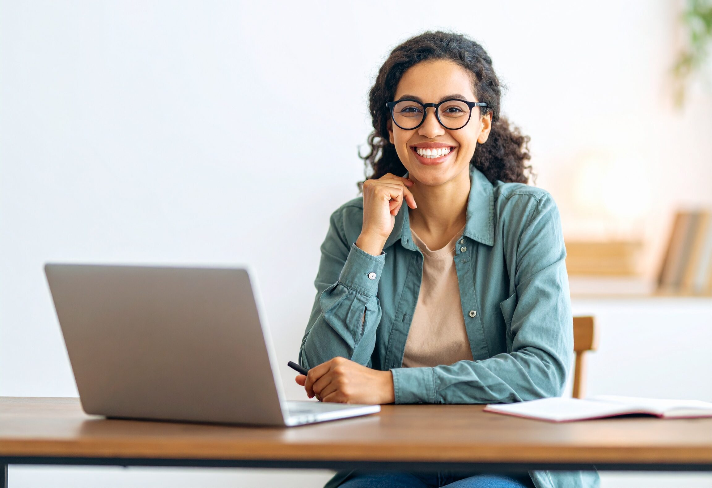 A smiling woman with glasses sitting at a desk, learning online with a laptop and notebook – promoting LINC Online English classes.