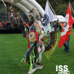An Indigenous dancer in full regalia — wearing a wolf headdress, colourful ribbon skirt with green and yellow fringe, eagle feather bustle, and hand-painted bag — stands on a grass field holding a ceremonial staff with feathers. A white First Nations flag and a Canadian flag are visible behind them, with a stage and orange-shirted participants in the background. The ISS of BC 'Welcoming Newcomers' logo appears in the bottom right corner.