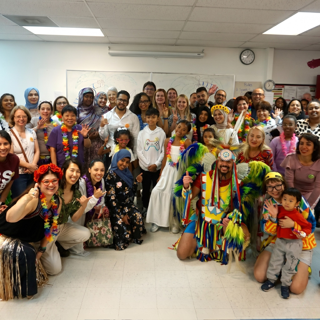 A large, diverse group of smiling people posing for a photo indoors, many wearing colorful traditional or cultural attire, including vibrant Indigenous regalia.
